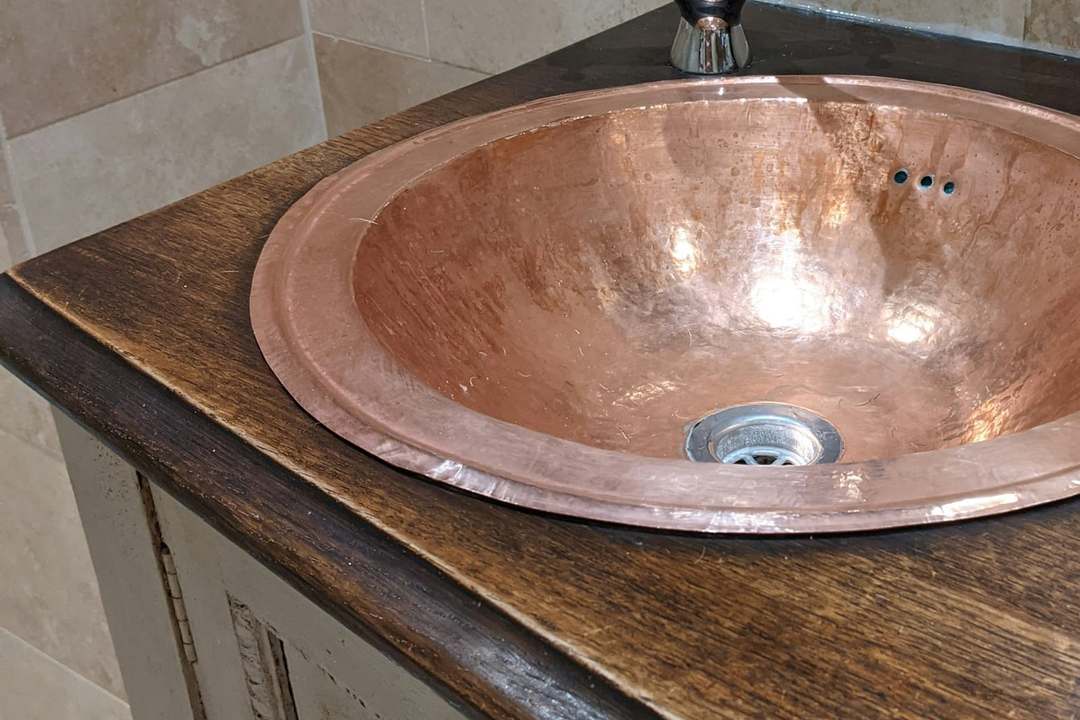Hammered copper basin set into a dark oak top on a carved painted cabinet, with travertine tiles behind