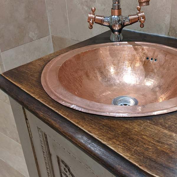 Hammered copper basin set into a dark oak top on a carved painted cabinet, with travertine tiles behind